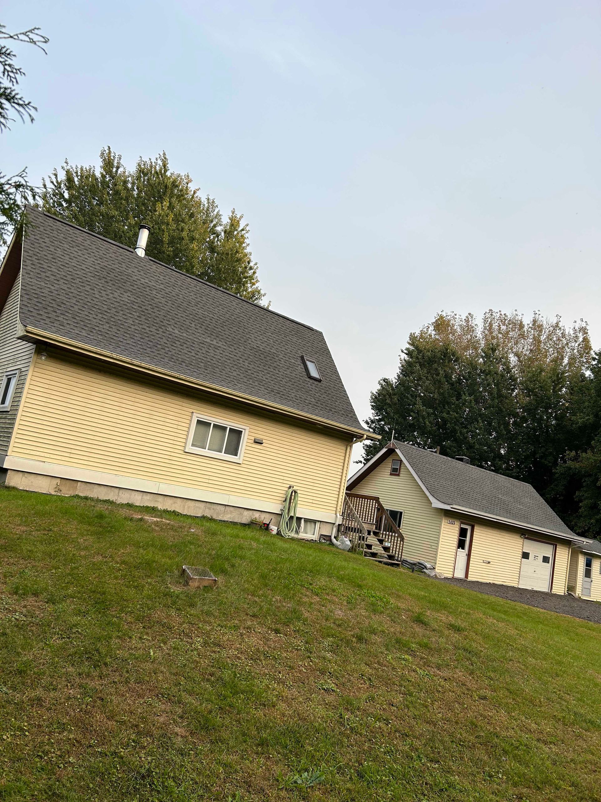 Maison jaune et garage indépendant sur une colline herbeuse, arbres en arrière-plan, sous un ciel nuageux.
