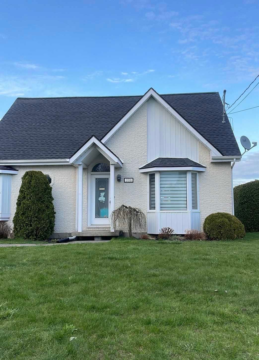 Maison en briques claires avec toit noir, petits buissons, pelouse verte et ciel bleu.