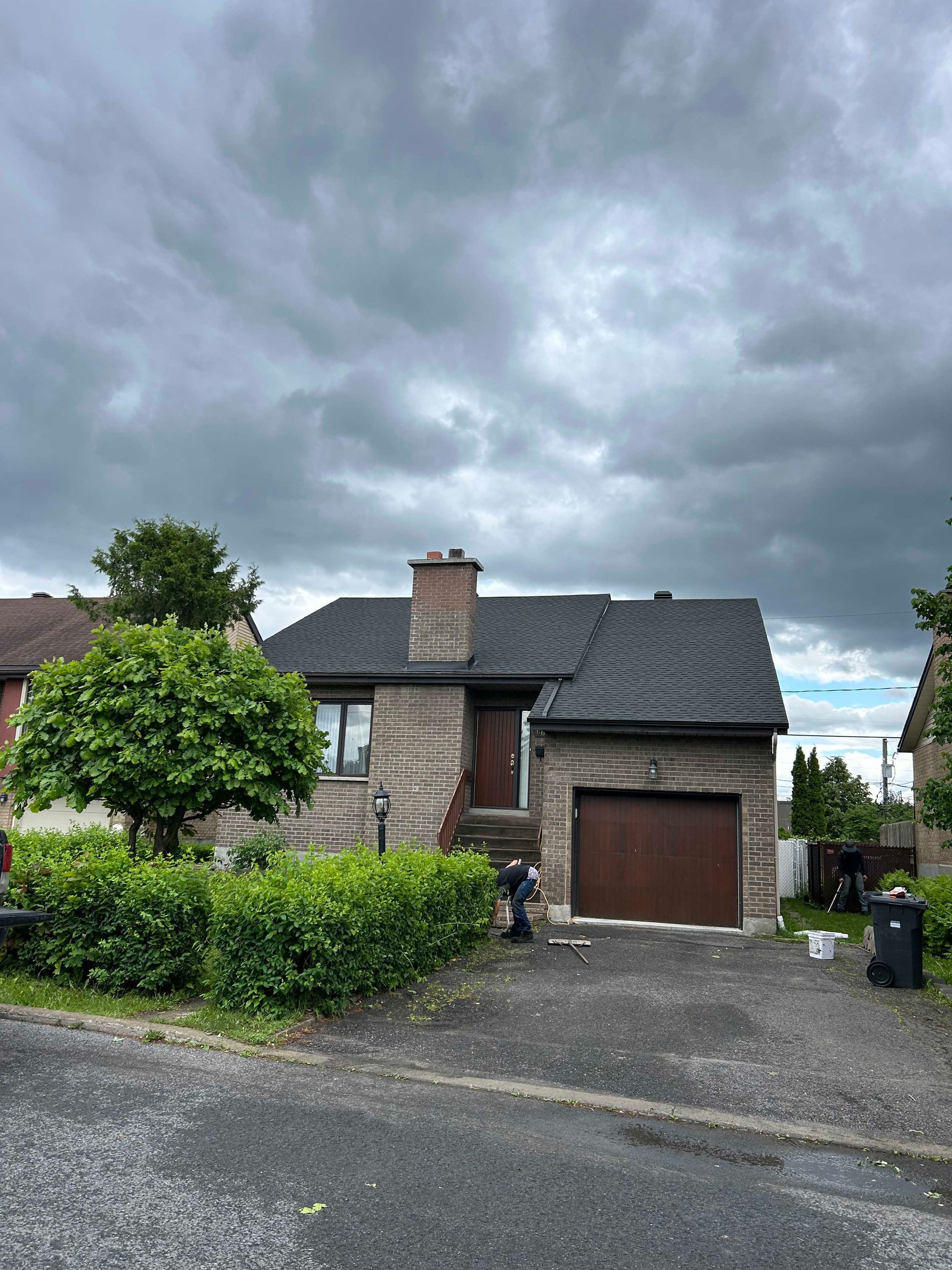Maison en briques au toit sombre et à la porte de garage marron fermée, sous un ciel nuageux.