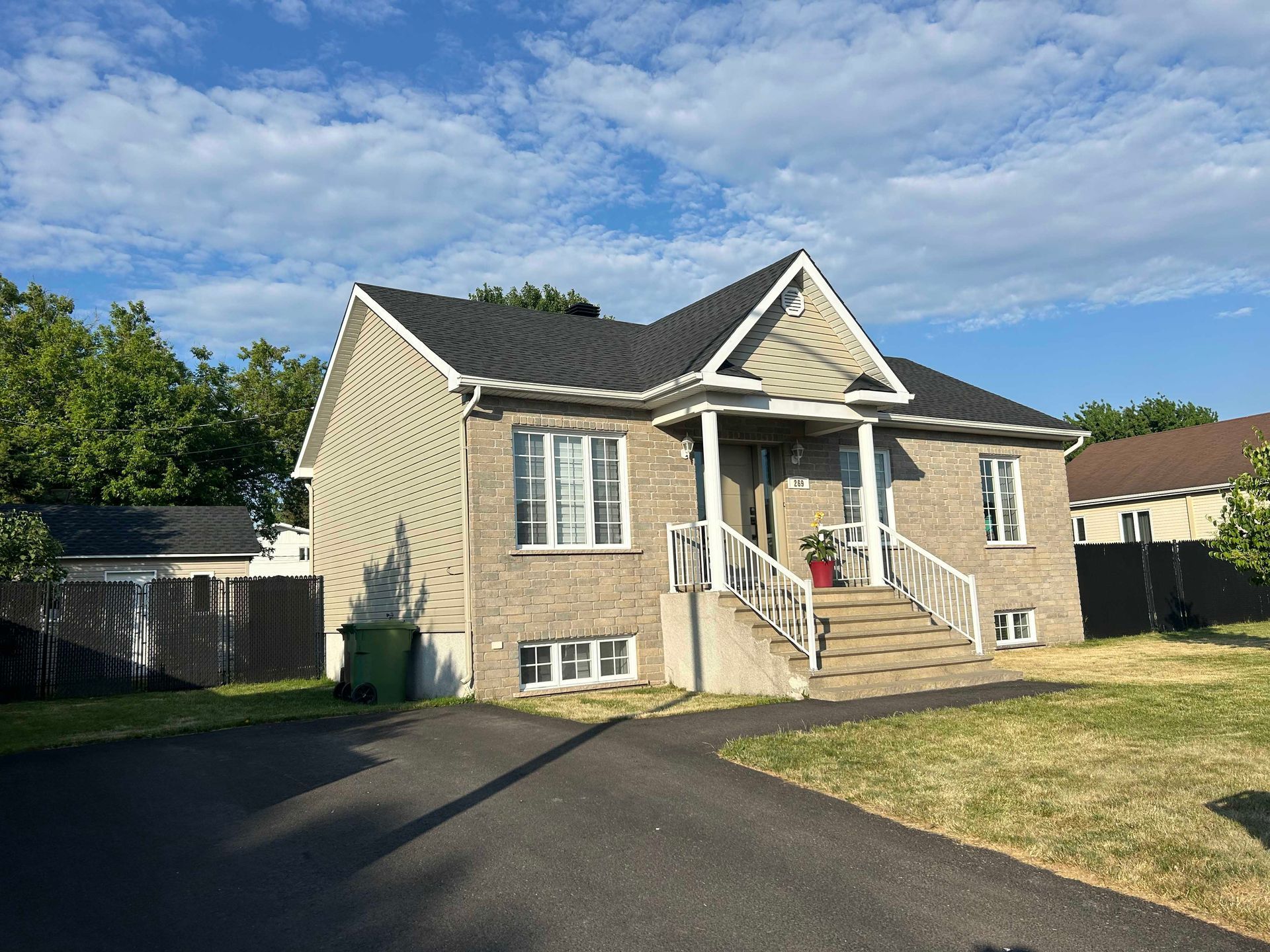 Maison en briques beiges avec toit noir, porche et allée, par une journée ensoleillée.