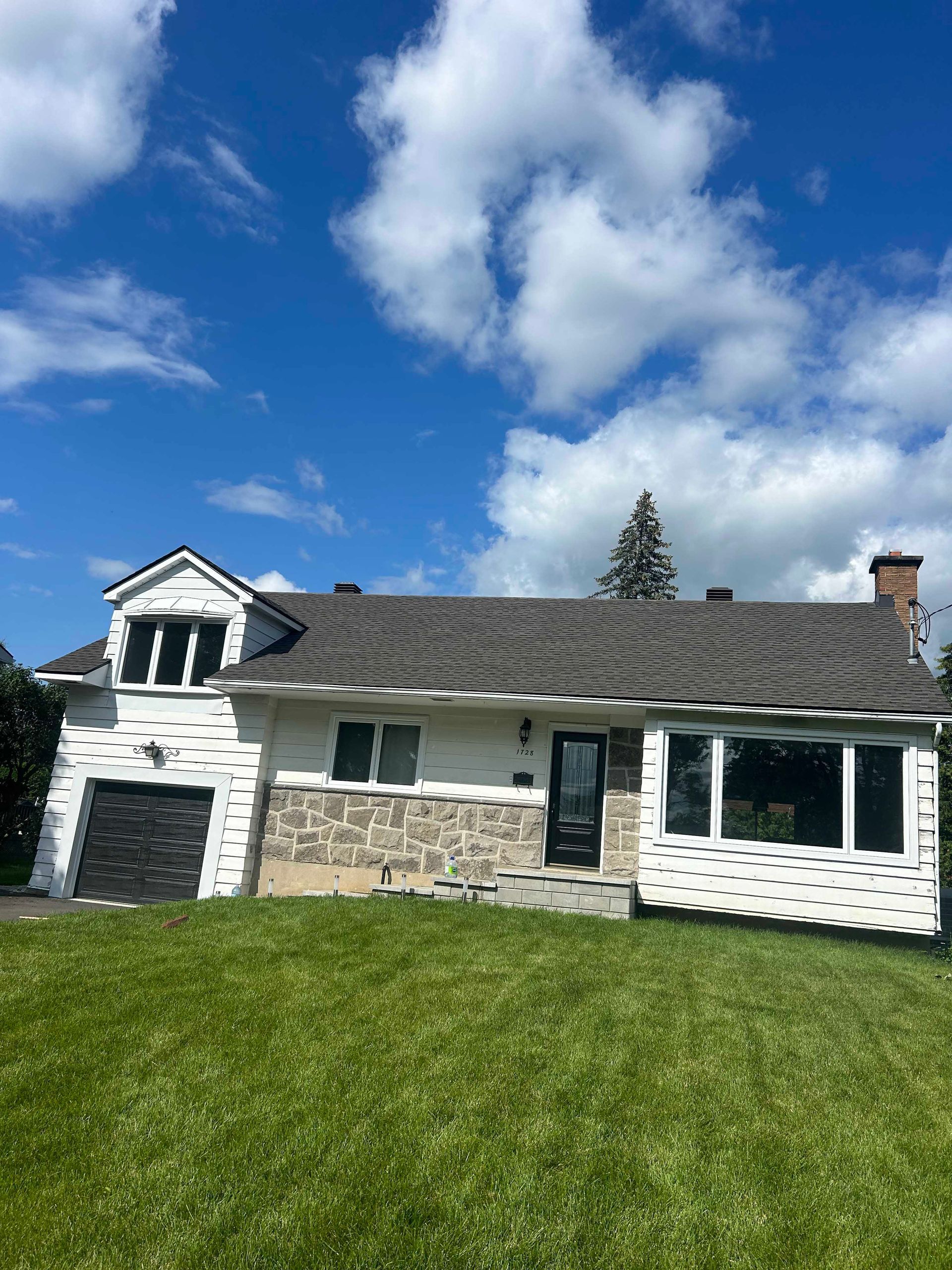 Maison à bardage blanc, façade en pierre et pelouse verte sous un ciel bleu nuageux.