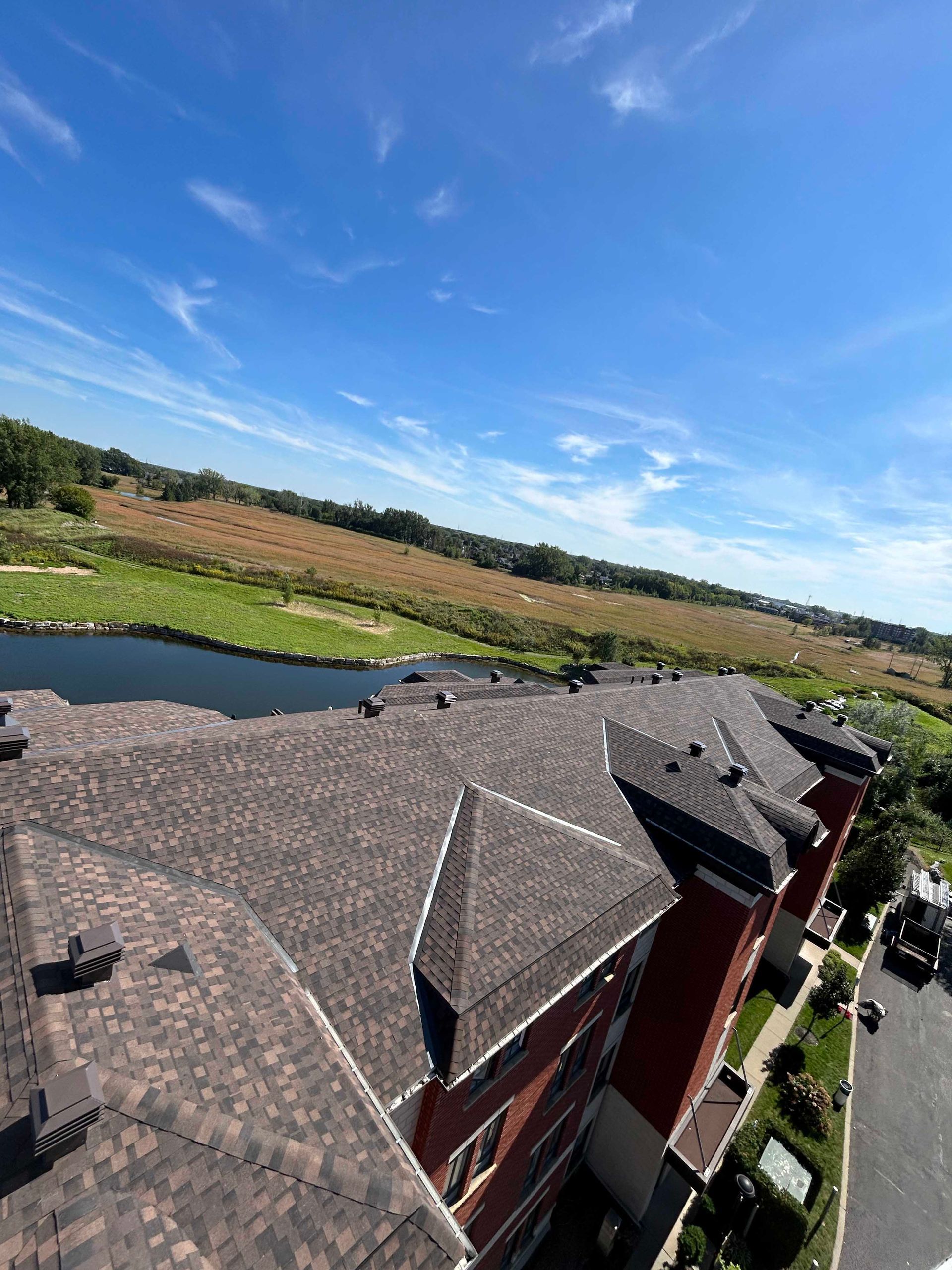 Vue depuis le toit d'un bâtiment au toit brun, surplombant un champ herbeux et un lac sous un ciel bleu nuageux.