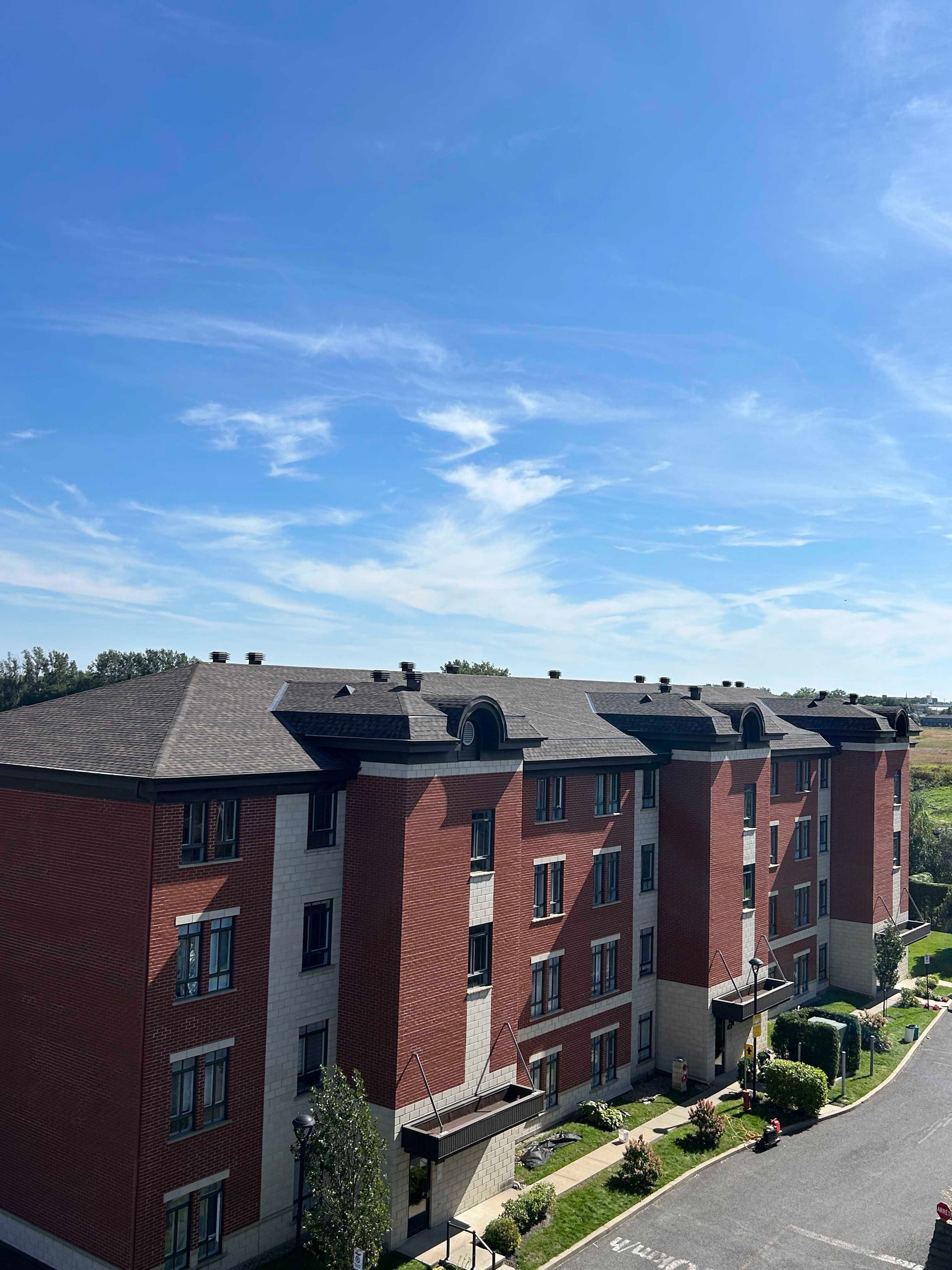 Immeuble d'appartements en briques rouges au toit sombre, sous un ciel bleu parsemé de nuages ​​vaporeux.