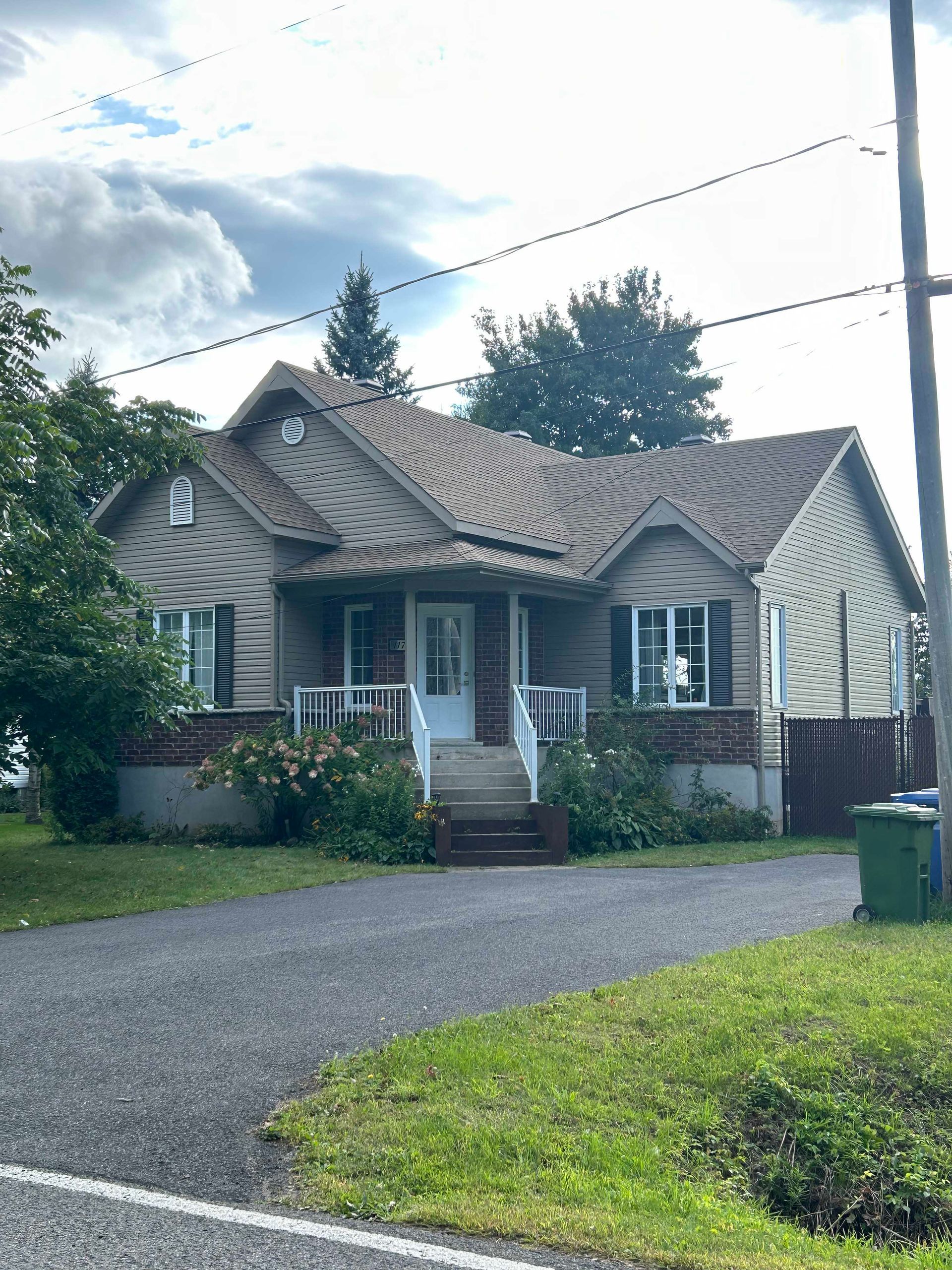 Maison aux murs beiges, au toit marron et aux fondations en briques rouges, avec porte blanche et porche, entourée d'une pelouse verte et d'une allée.