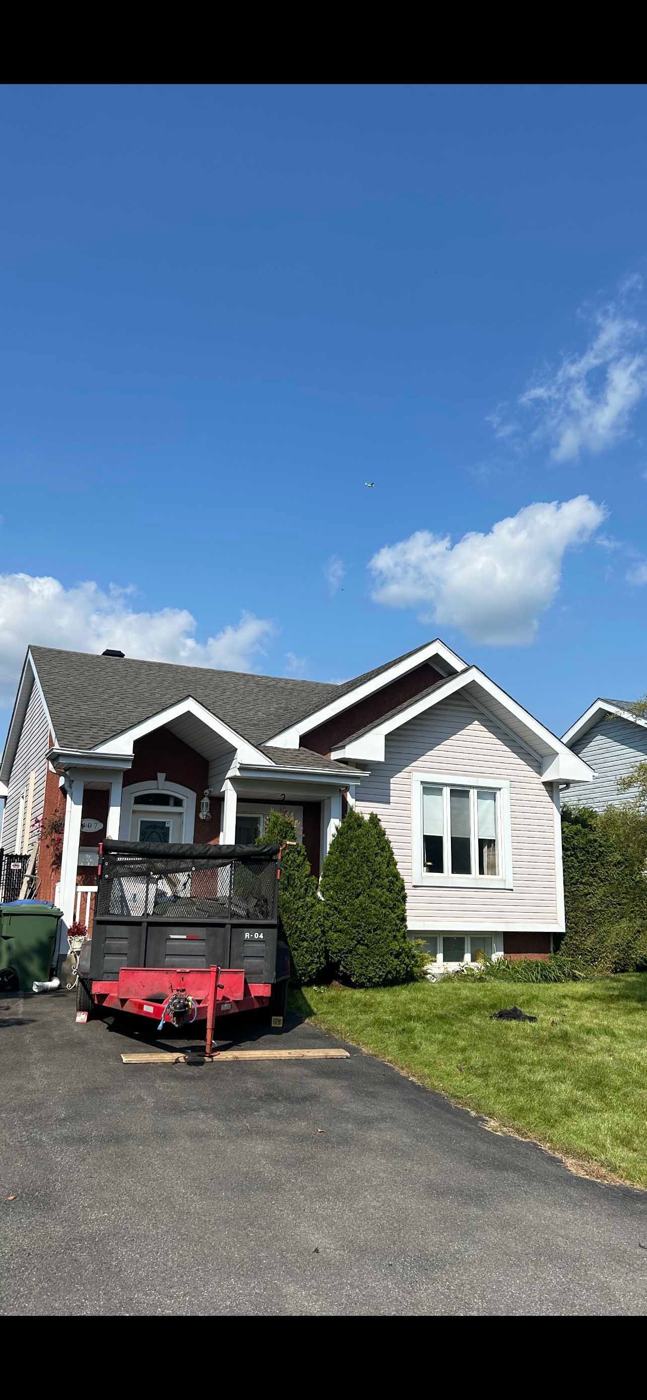 Une maison rose pâle avec un toit gris et un ciel bleu. Une caravane rouge est garée dans l'allée.