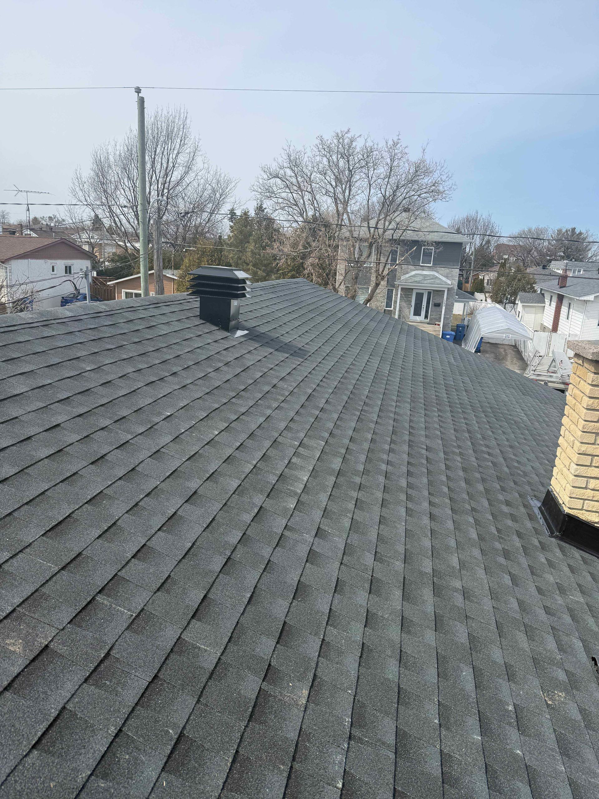 Toit en bardeaux d'asphalte foncé avec cheminée et ventilation, sur fond d'arbres, de bâtiments et d'un ciel lumineux.