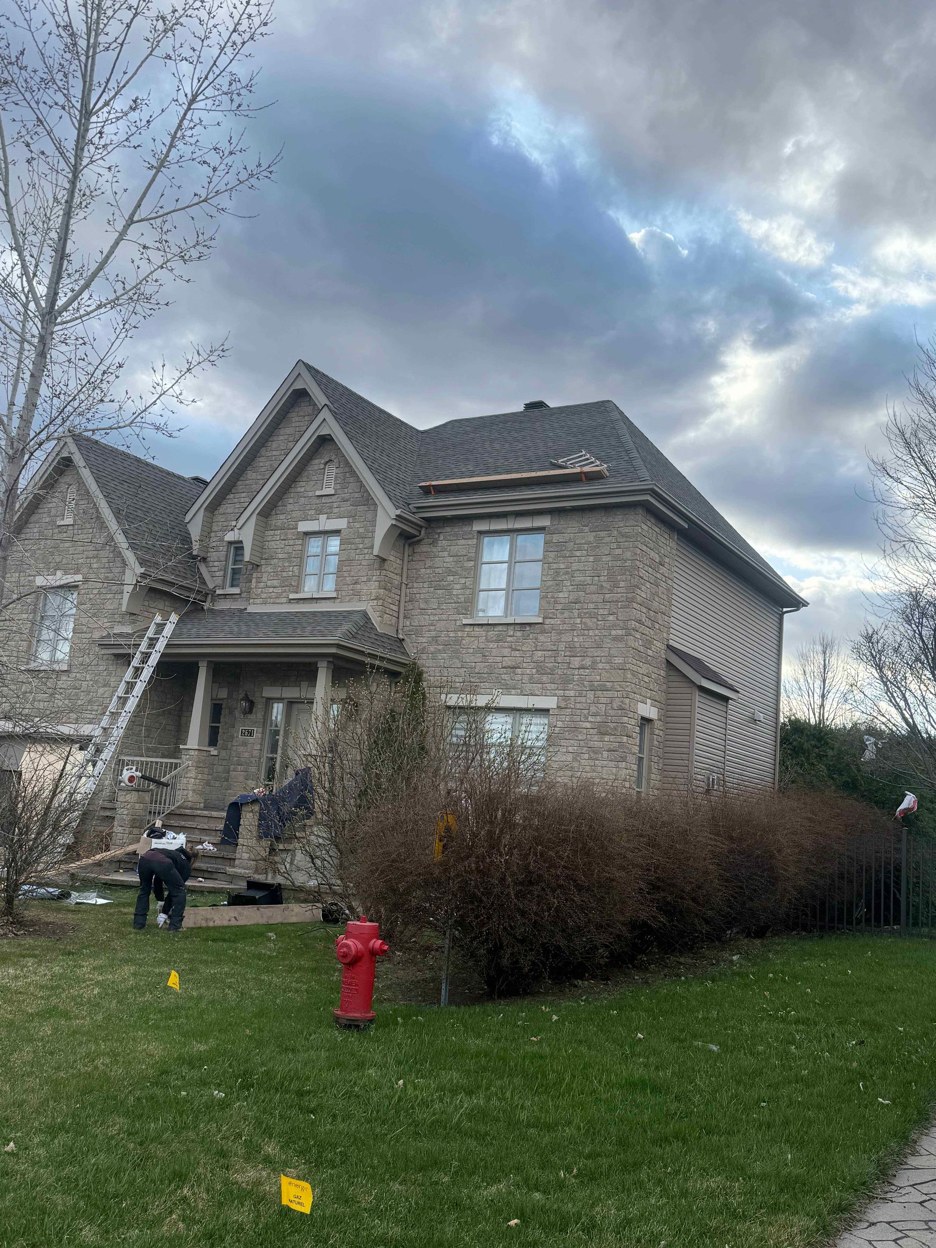 Maison en pierre à deux étages avec toit sombre ; ouvriers sur une échelle ; ciel nuageux, pelouse verte et borne d'incendie rouge devant.