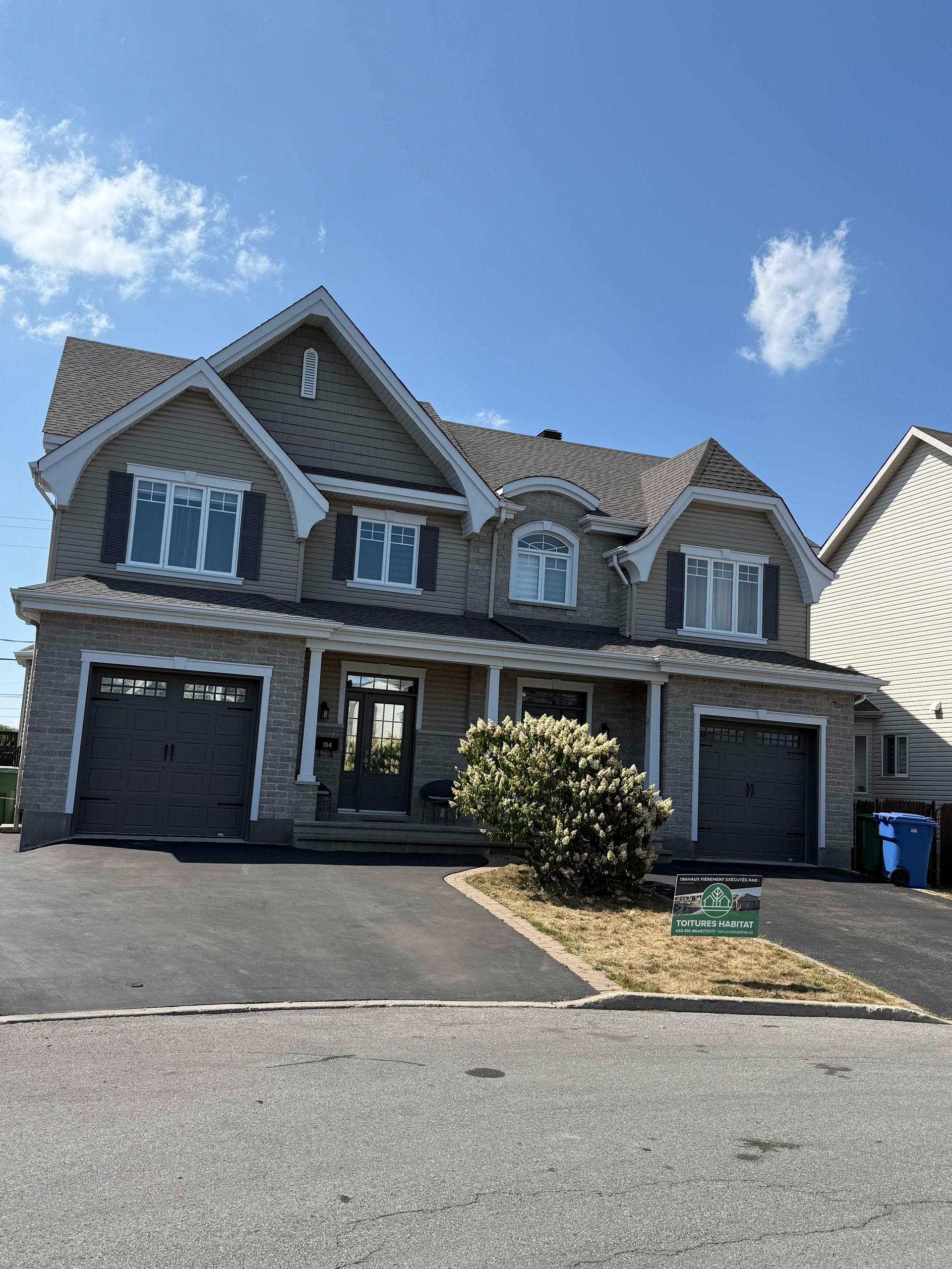 Maison à deux étages avec façade en pierre grise et en brique, deux portes de garage et une cour avant sous un ciel bleu.