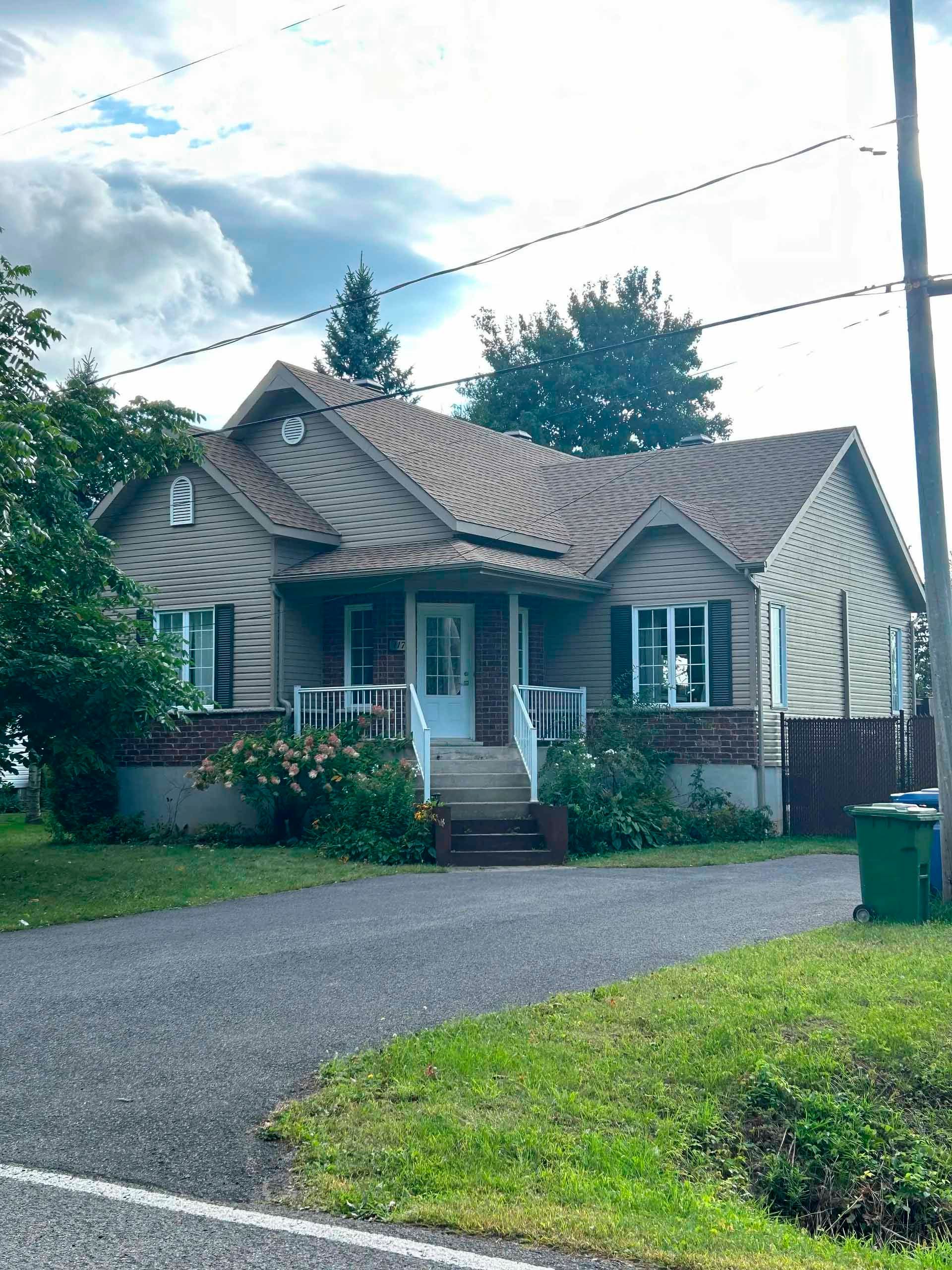 Maison avec toit marron, bardage beige, soubassement en briques rouges, porte blanche et allée en gravier.