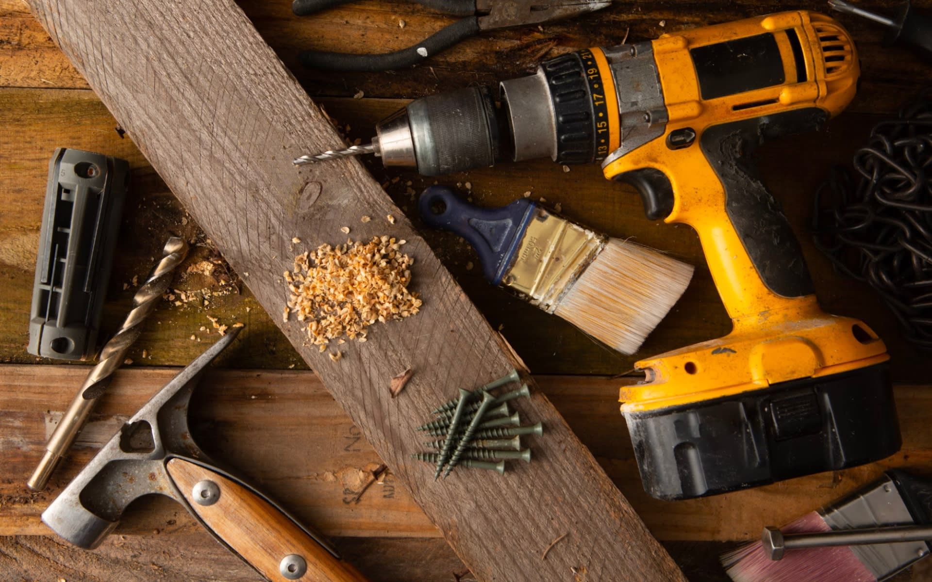 A drill , hammer , brush , screws and other tools are on a wooden table.