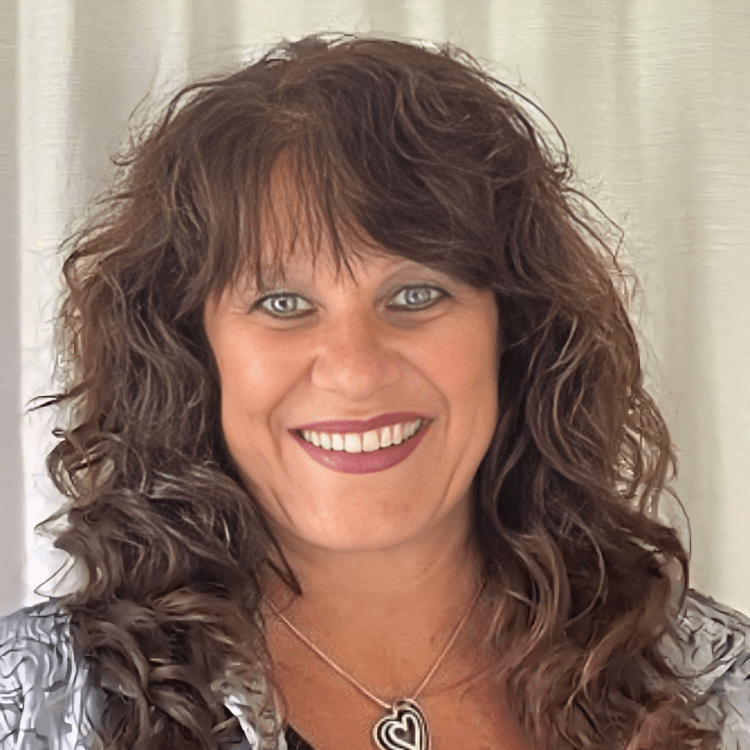 A woman with curly hair is smiling and wearing a necklace with a heart pendant.
