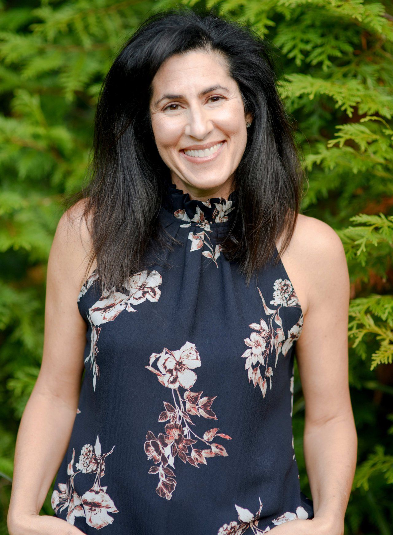 A woman in a floral dress is smiling in front of a tree.