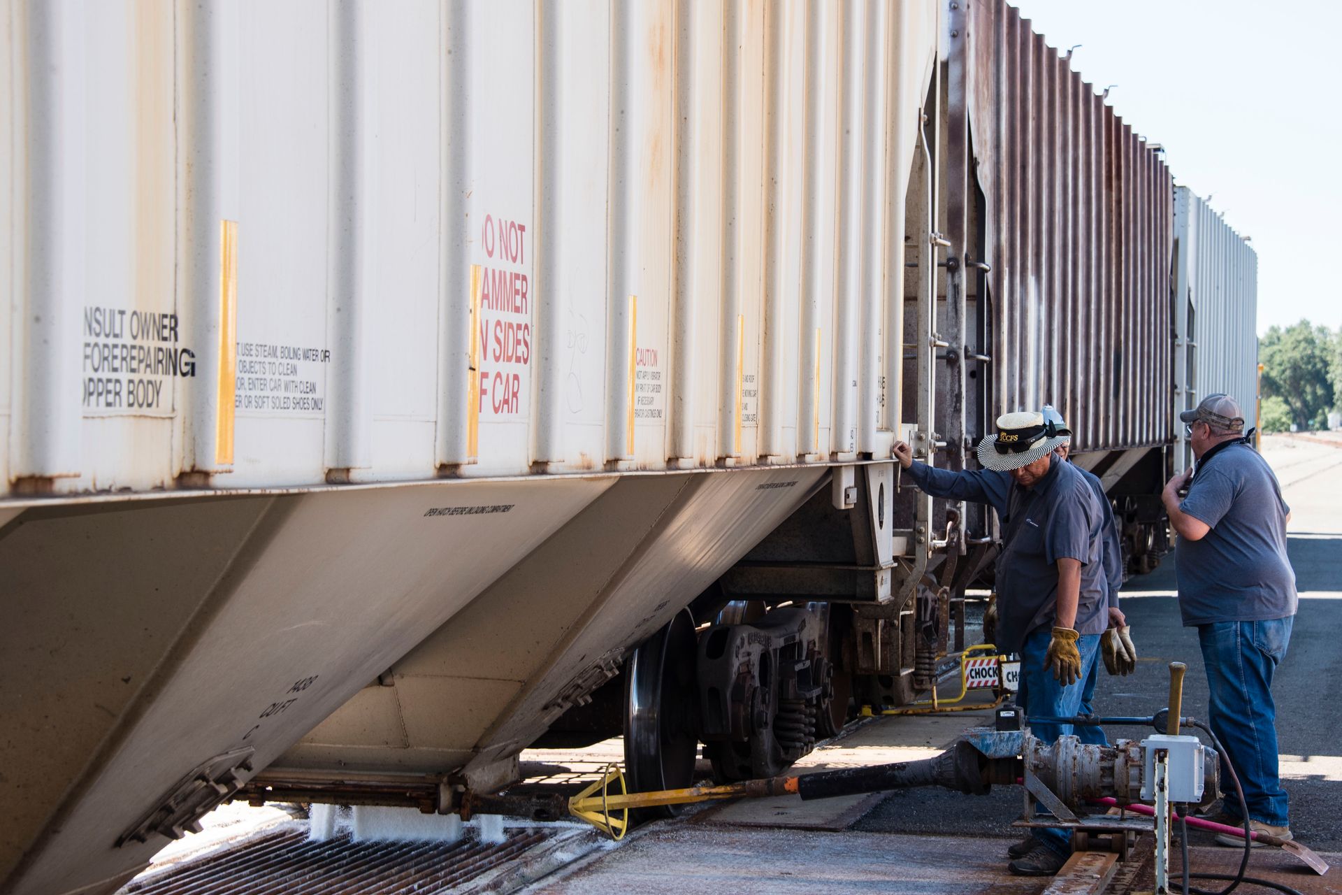 Two people inspect a tilted white freight train car, outdoors.