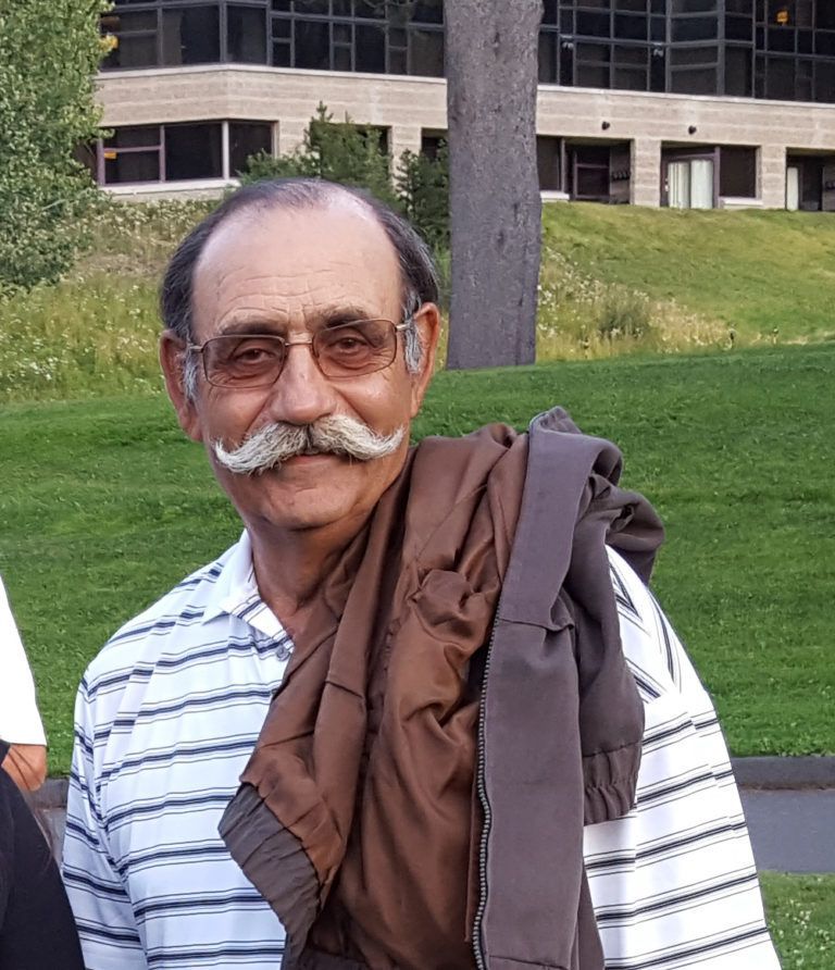 Man with a large mustache and glasses, holding a brown jacket over his shoulder, smiling outside.