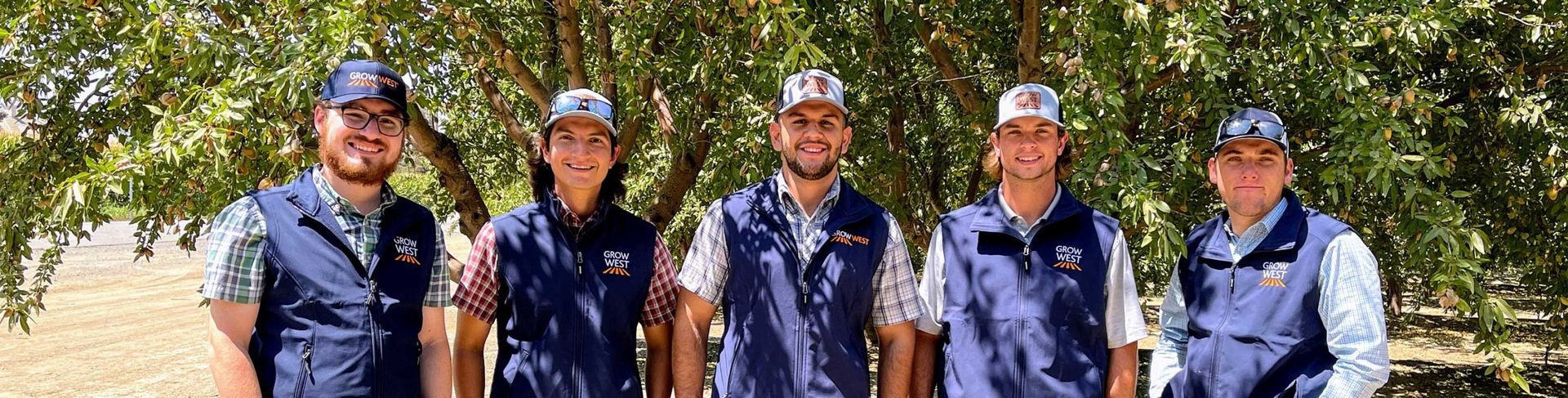 Five people in navy vests and caps standing outdoors under trees.
