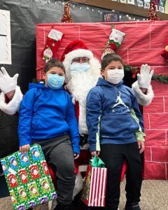 Two children in masks pose with Santa Claus; holiday gifts in hand. Festive backdrop with stockings.