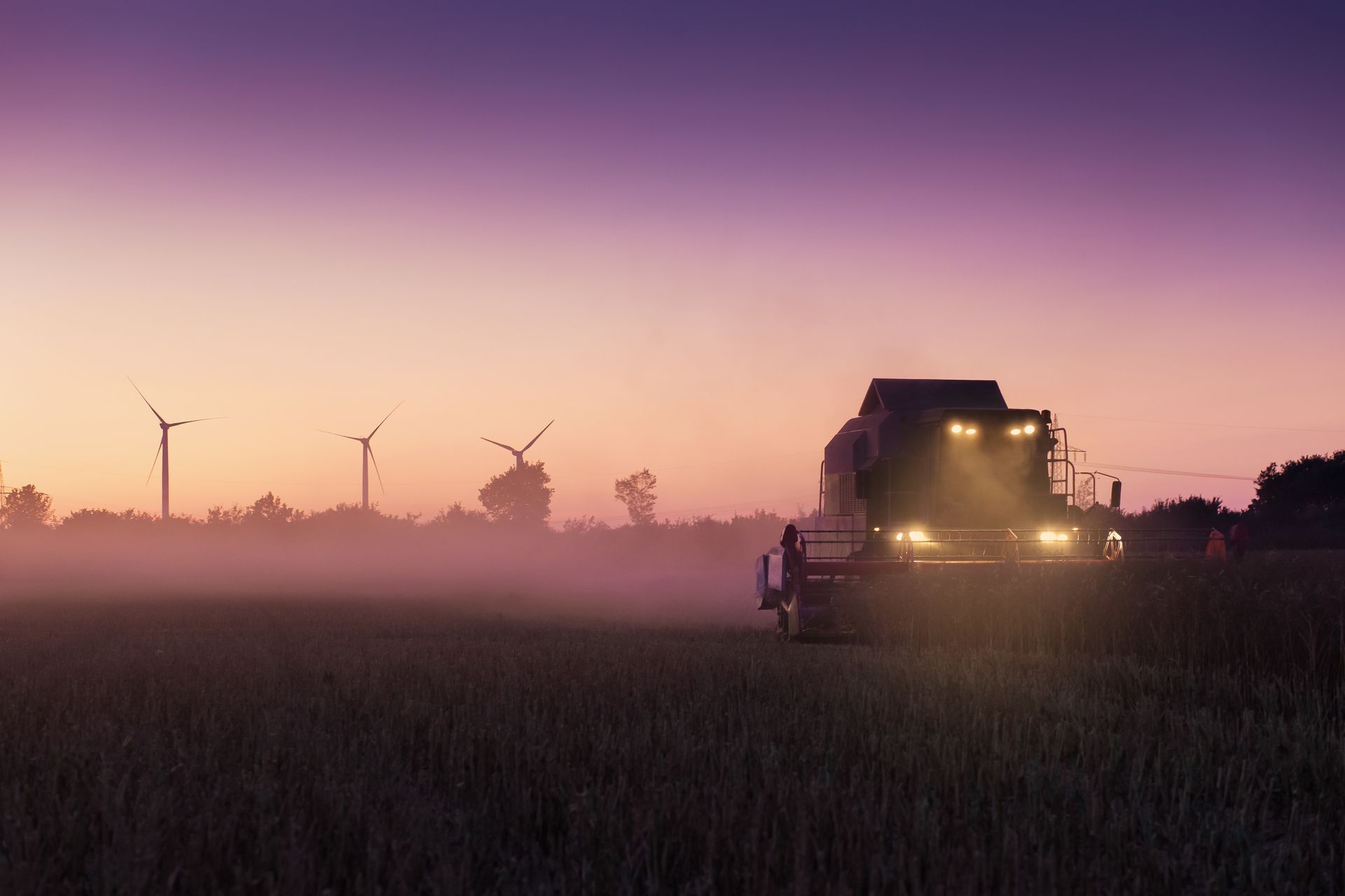Combine harvester in a field at dusk, with windmills in the background and a purple sky.