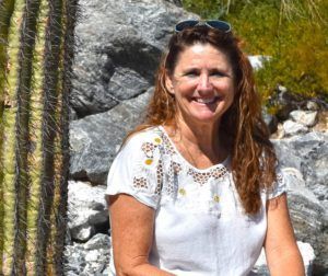 Woman with long hair smiles, wearing a white embroidered top, with sunglasses on her head; sitting outside near a cactus.