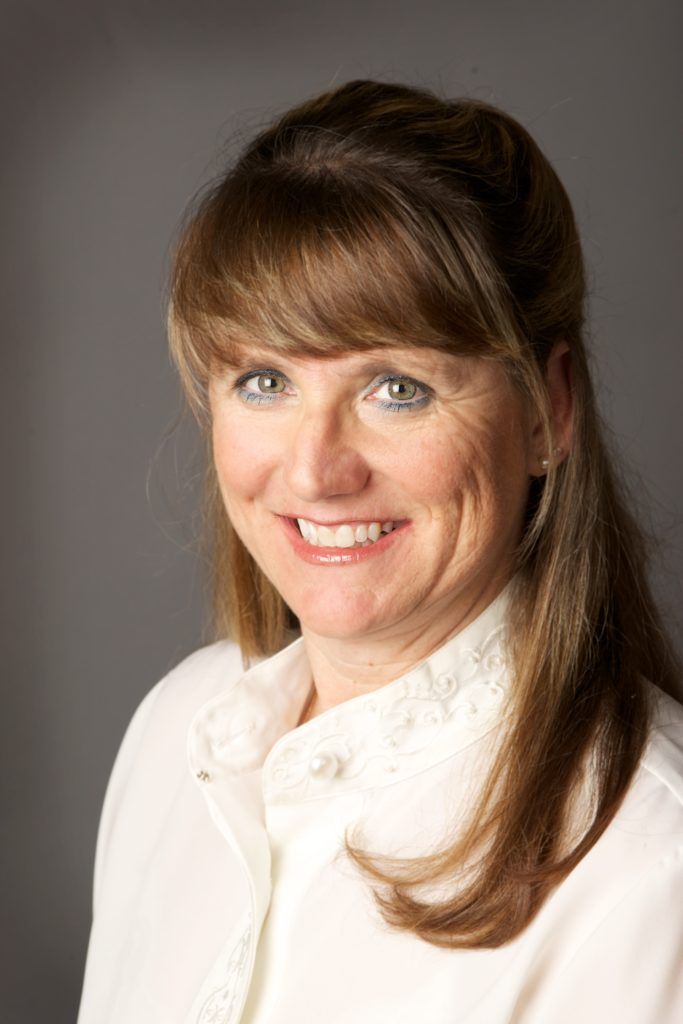 Woman with light brown hair smiles, wearing a white shirt against a gray background.