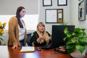 Two women at desk looking at computer; one points, other watches. Office setting with plants.