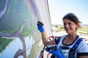 Woman in a lift paints a mural on a building, holding a brush and paint bucket, smiling.