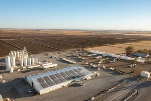 Aerial view of agricultural site: fields, buildings, storage tanks, and transportation vehicles under a clear sky.