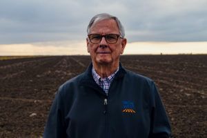 Man with glasses stands in a tilled field, wearing a navy jacket. Overcast sky in background.