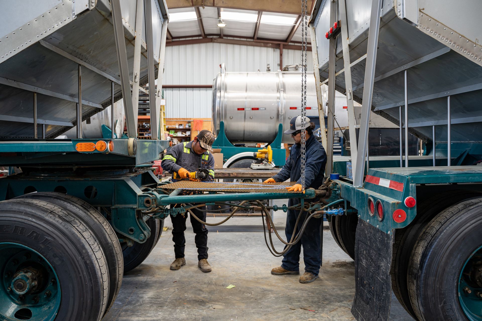 Two workers repair a truck trailer in a garage; one grinds metal, the other inspects the work.