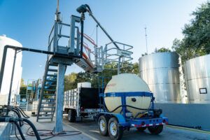 A truck with a tank trailer being loaded with a liquid, using a lifting platform with a conveyor, near storage tanks.