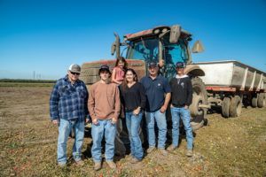 Family poses in front of a tractor and trailer in a field on a sunny day.