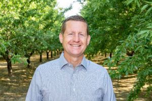 Man smiling in an orchard, wearing a light blue button-down shirt. Green trees in background.