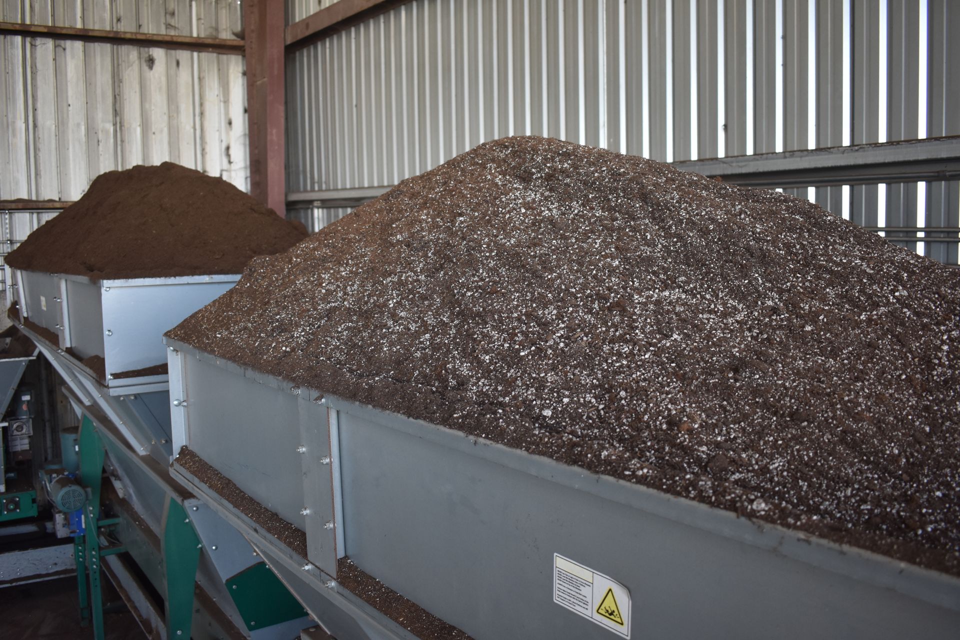Two large bins overflowing with dark, granular material on a conveyor belt, indoors.