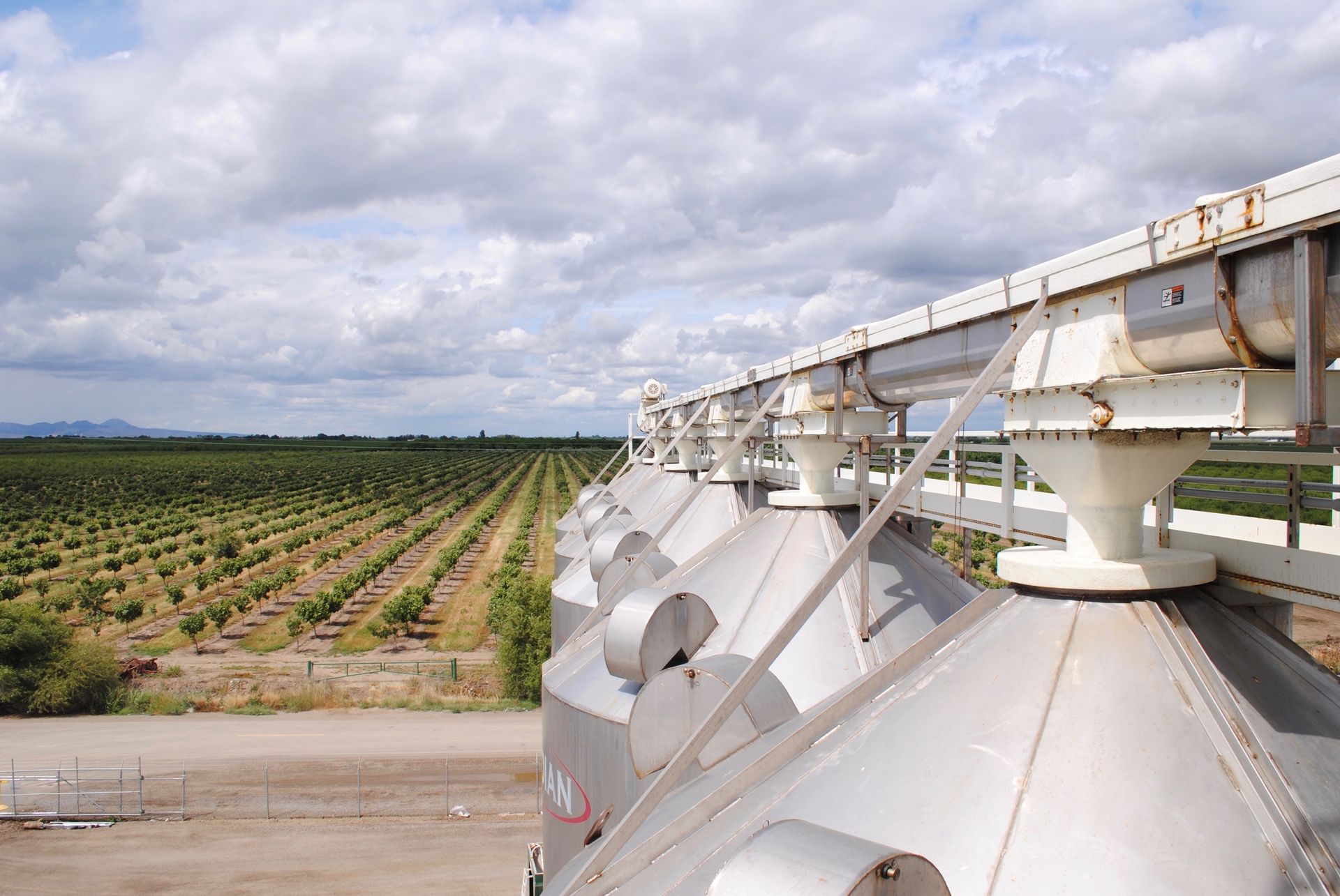 Elevated view of industrial grain silos with conveyor belt, overlooking a large orchard under cloudy sky.