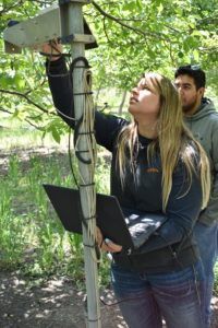Woman adjusting sensor on pole, holding laptop. Man looks on in a wooded area.