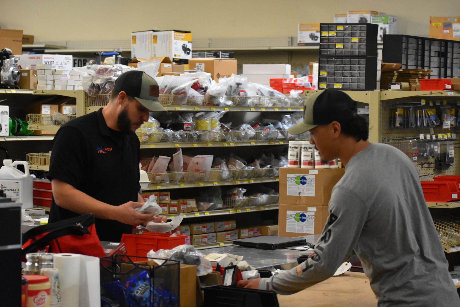 Two men in a store: one at a counter, sorting items, the other looking at a box. Shelves stocked with products.