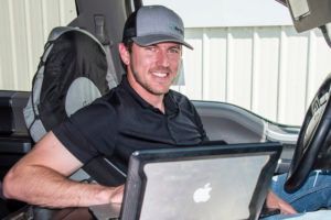 Man in truck cab using laptop, smiling, wearing cap and black shirt.