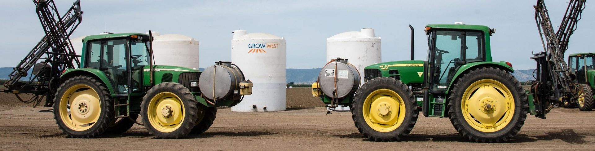 Two green tractors with yellow wheels in a field; tall white tanks in the background.