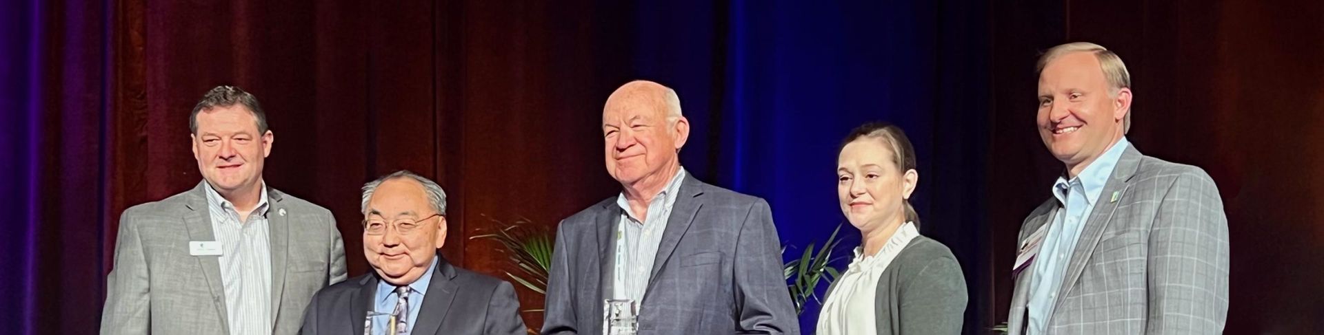 Five people stand in front of a stage curtain. They are all wearing business attire and smiling.