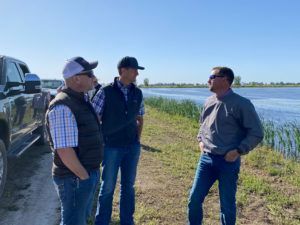 Three men in casual wear standing near a body of water, possibly discussing something.