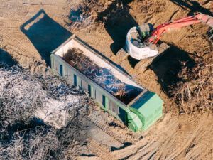 Green dumpster being filled by a white excavator at a construction site.