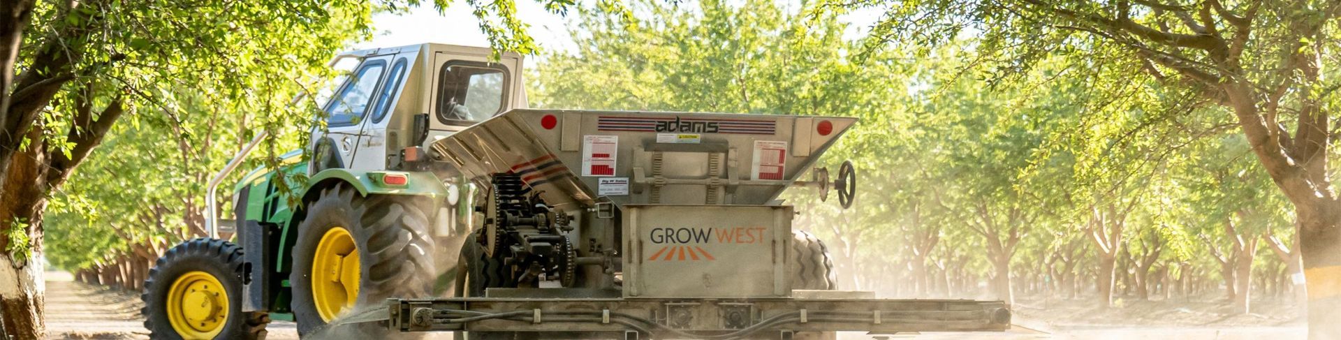 A tractor spreading fertilizer in an orchard, with bright green foliage in the background.