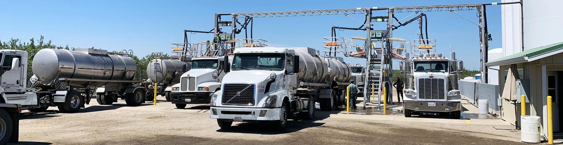Tanker trucks parked at a facility under a blue sky, with metal framework overhead.