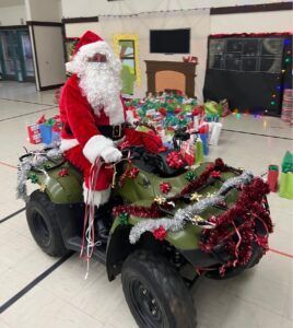 Santa Claus on an ATV decorated with Christmas garland, surrounded by presents in a gymnasium.