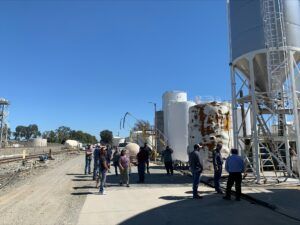 Group of people standing near industrial tanks outdoors on a sunny day.