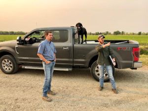 Two men and a dog by a pickup truck in a field, one man points.