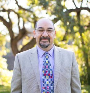 Man in light suit, lavender shirt, patterned tie, glasses, outdoors, smiling in front of a tree.