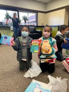 Two children kneeling, holding gifts, wearing masks, in a room. A woman stands in the background.