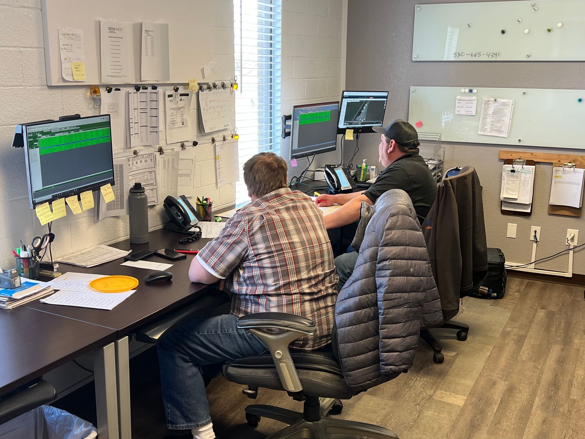 Two people at desks with computers, documents, and whiteboard in an office setting.