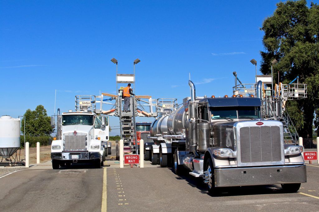Trucks at a facility, likely for liquid transfer. Man on platform, blue sky, concrete ground.