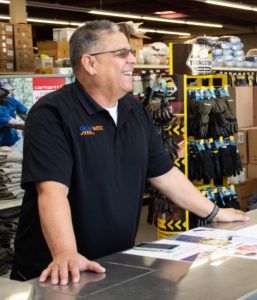 Man in black shirt smiles at counter in store. Shelves of merchandise in background.